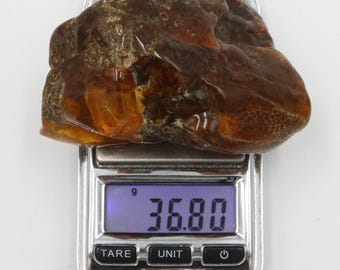 May include: A rough, amber-colored stone rests on a digital scale. The scale displays a weight of 36.80 grams. The stone has a textured surface with areas of translucence, showcasing its natural, organic form. The scale is silver with black buttons.