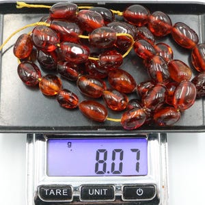 May include: A necklace of polished, oval amber beads, strung on a yellow cord, presented on a black tray. The beads are a rich, translucent, reddish-brown colour. The digital scale below reads 8.07 grams.