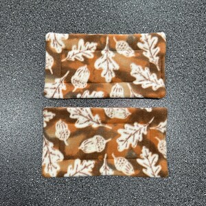 May include: Two rectangular pet bed pads. The pads feature a brown and tan background with a repeating pattern of white oak leaves and acorns. The edges are a darker brown color.