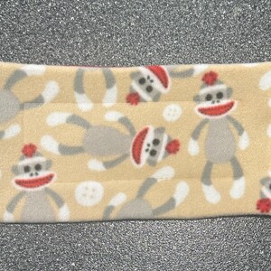 May include: A beige fabric rectangle with a repeating pattern of gray monkeys wearing red and white hats. The monkeys are in various poses, some with their arms raised and some with their arms down.