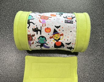 Könnte beinhalten: Ein hellgrünes Fleece-Haustierbett mit einem passenden rechteckigen Pad. Das zylindrische Bett hat einen weißen Stoff mit einem Halloween-Motivdruck von Cartoon-Charakteren, Kürbissen, Fledermäusen und Konfetti.