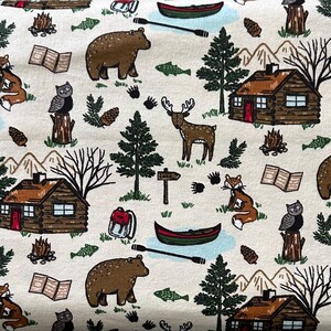 May include: A cream-colored fabric featuring a repeating pattern of woodland and camping elements. The design includes brown bears, deer, foxes, owls, canoes, cabins, trees, and campfires. The fabric is suitable for various craft projects.