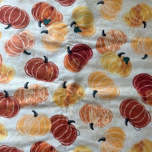 May include: A cream-colored fabric featuring a repeating pattern of pumpkins in various shades of orange, yellow, and deep red. Each pumpkin is illustrated with visible line details and small green stems, distributed evenly across the textile surface.
