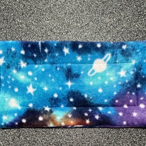 May include: A blue and purple fleece pet bed with a galaxy print featuring stars and a planet. The bed is quilted and has a white border.