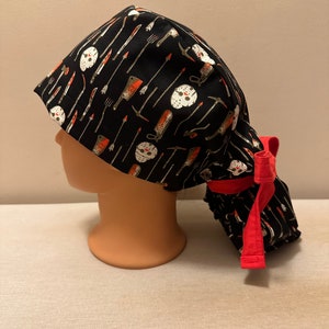 K&ouml;nnte beinhalten: Schwarze OP-M&uuml;tze mit roter Schleife. Die M&uuml;tze hat ein Muster aus wei&szlig;en Hockeymasken und verschiedenen Waffen, darunter Messer, &Auml;xte und eine Machete.