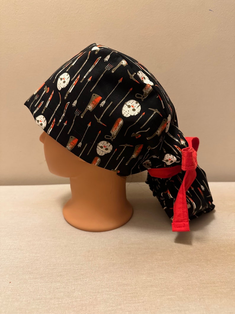 K&ouml;nnte beinhalten: Schwarze OP-M&uuml;tze mit roter Schleife. Die M&uuml;tze hat ein Muster aus wei&szlig;en Hockeymasken und verschiedenen Waffen, darunter Messer, &Auml;xte und eine Machete.
