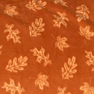 May include: A fabric with a burnt orange background featuring a repeating pattern of light beige oak leaves. The leaves are oriented in various directions across the surface, creating a consistent and uniform design throughout the textile material.