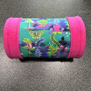 May include: A cylindrical pillow with a vibrant frog and butterfly print on a teal background. The ends are a bright pink fleece. The design features colorful illustrations of frogs, butterflies, and foliage, creating a whimsical and playful aesthetic.