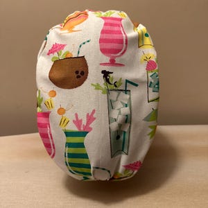 May include: White fabric scrub hat with a tropical cocktail print featuring pink, green, yellow, and brown colors.