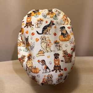 May include: A white fabric scrub hat with a pattern of dogs and pumpkins. The dogs are wearing fall-themed outfits and some have pumpkins on their heads. The text "Oh, Fall!" is visible on the hat.