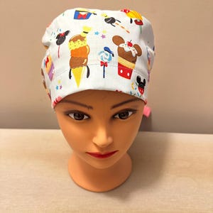 May include: White scrub hat with a colourful print of Mickey Mouse and Disney treats, including ice cream cones, cupcakes and candy.