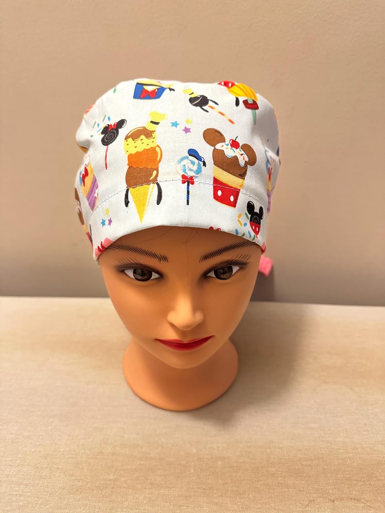 May include: White scrub hat with a colourful print of Mickey Mouse and Disney treats, including ice cream cones, cupcakes and candy.