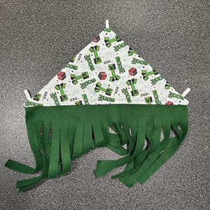 May include: A triangular pet bed with a white fabric top featuring a green Creeper and red cube pattern. The bed has a green fringe along the bottom edge and white support rods.