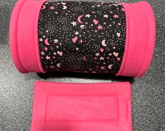 Könnte beinhalten: Ein rosa-schwarzes Fleece-Tierbett in Zylinderform. Der Zylinder hat einen schwarzen Stoff mit einem rosa-weißen Muster aus Sternen, Monden, Herzen und Sternschnuppen. Zum Bett gehört ein flaches rosa Fleece-Kissen.