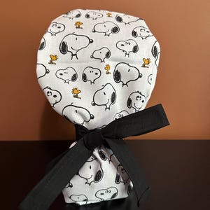 May include: A white scrub cap featuring a repeating pattern of black and white Snoopy illustrations and small yellow Woodstock figures. The cap includes a solid black fabric tie at the back, which is knotted to secure the gathered fabric in place.