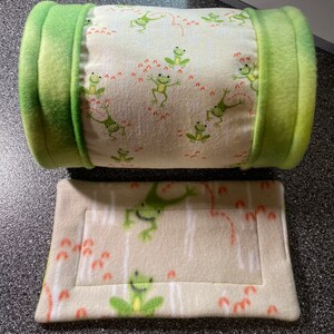 May include: A green and white fleece pet bed set. The cylindrical bed has green ends and a cream-colored center with green frog illustrations and orange accents. A matching rectangular pad is below.