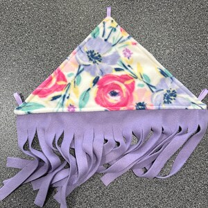 May include: A triangular fleece hammock featuring a floral pattern with pink roses, purple flowers, and green leaves on a cream background. The bottom edge has a solid light purple fringe, and small fabric loops are attached to the three corners.