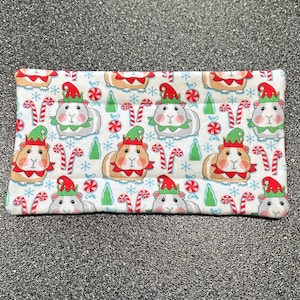 May include: A rectangular, white fabric item with a repeating pattern of guinea pigs wearing elf hats, candy canes, Christmas trees, and red and white peppermint candies. The guinea pigs have brown, white, and gray fur.