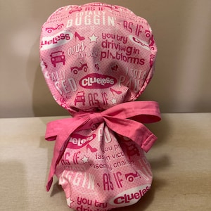May include: Pink fabric scrub hat with a white print of the words "Clueless" and "As If" repeated in a pattern. The hat is tied with a pink ribbon.