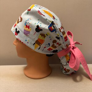 May include: A light blue surgical scrub hat with a pink tie. The hat features a pattern of cartoon characters and desserts, including Mickey Mouse, Minnie Mouse, ice cream cones, cupcakes, and Mickey Mouse ears.