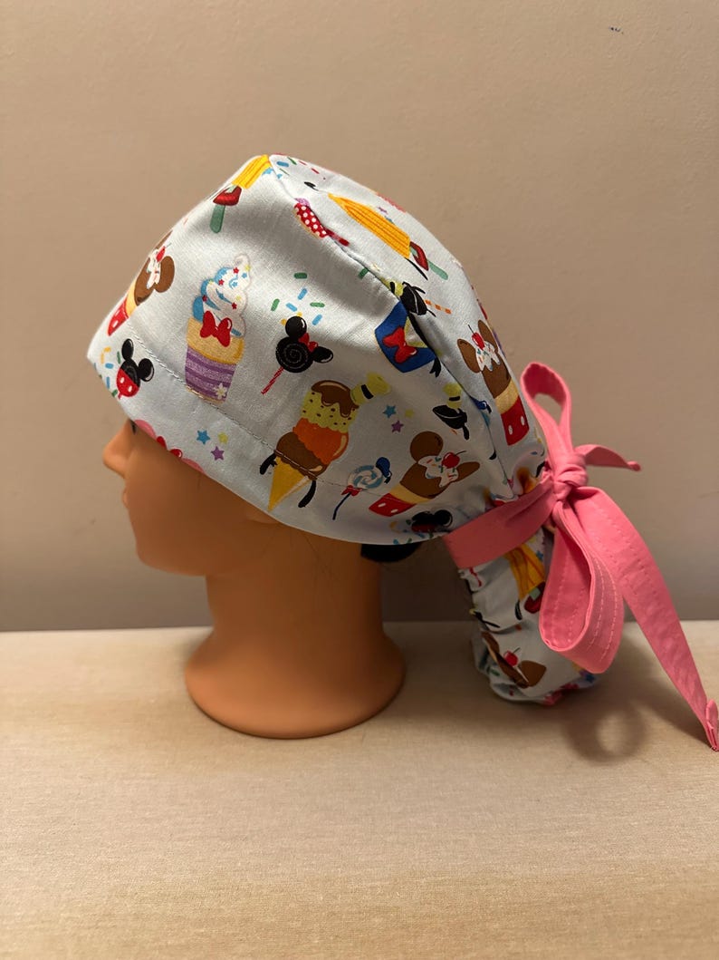 May include: A light blue surgical scrub hat with a pink tie. The hat features a pattern of cartoon characters and desserts, including Mickey Mouse, Minnie Mouse, ice cream cones, cupcakes, and Mickey Mouse ears.