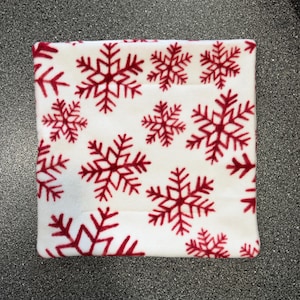 May include: A square fabric coaster featuring a white background with a repeating pattern of red snowflakes. The textile has a soft, plush texture and finished edges, displaying a consistent print of various sized snowflake shapes across the surface.