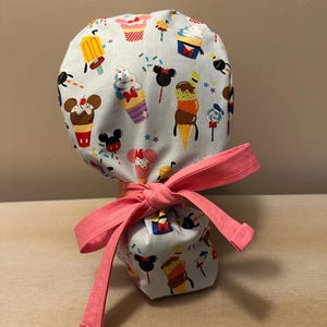 May include: A light blue scrub hat with a pink ribbon tied around it. The hat is decorated with a pattern of ice cream cones, cupcakes, and Mickey Mouse heads.