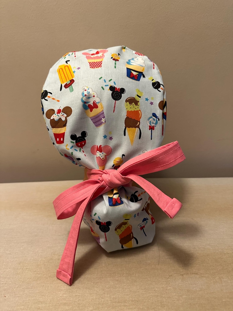 May include: A light blue scrub hat with a pink ribbon tied around it. The hat is decorated with a pattern of ice cream cones, cupcakes, and Mickey Mouse heads.