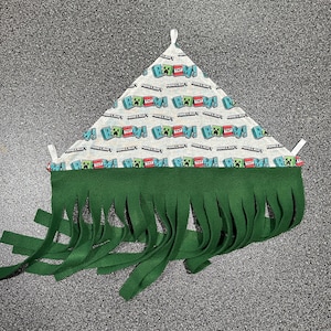 May include: A triangular pet bed with a Minecraft theme. The top is patterned with Minecraft characters and the word "TNT". The bottom edge features a green fringe.