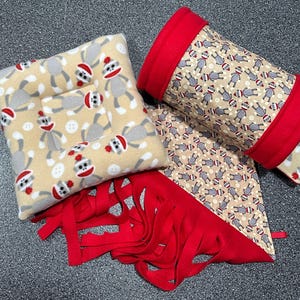 May include: A set of three plush toys featuring a monkey pattern. The set includes a pillow, a tube, and a blanket with fringe. The toys are beige with red accents and a repeating pattern of monkeys wearing red hats.