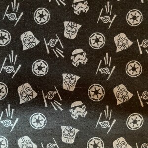 May include: Black fabric with a repeating pattern of white Star Wars characters and symbols. The characters include Darth Vader, Stormtroopers, and the Death Star symbol.