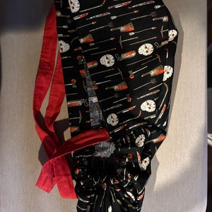 K&ouml;nnte beinhalten: Eine schwarze OP-M&uuml;tze mit roter Schleife. Die M&uuml;tze hat ein sich wiederholendes Muster aus wei&szlig;en Hockeymasken, Messern und anderen Waffen.