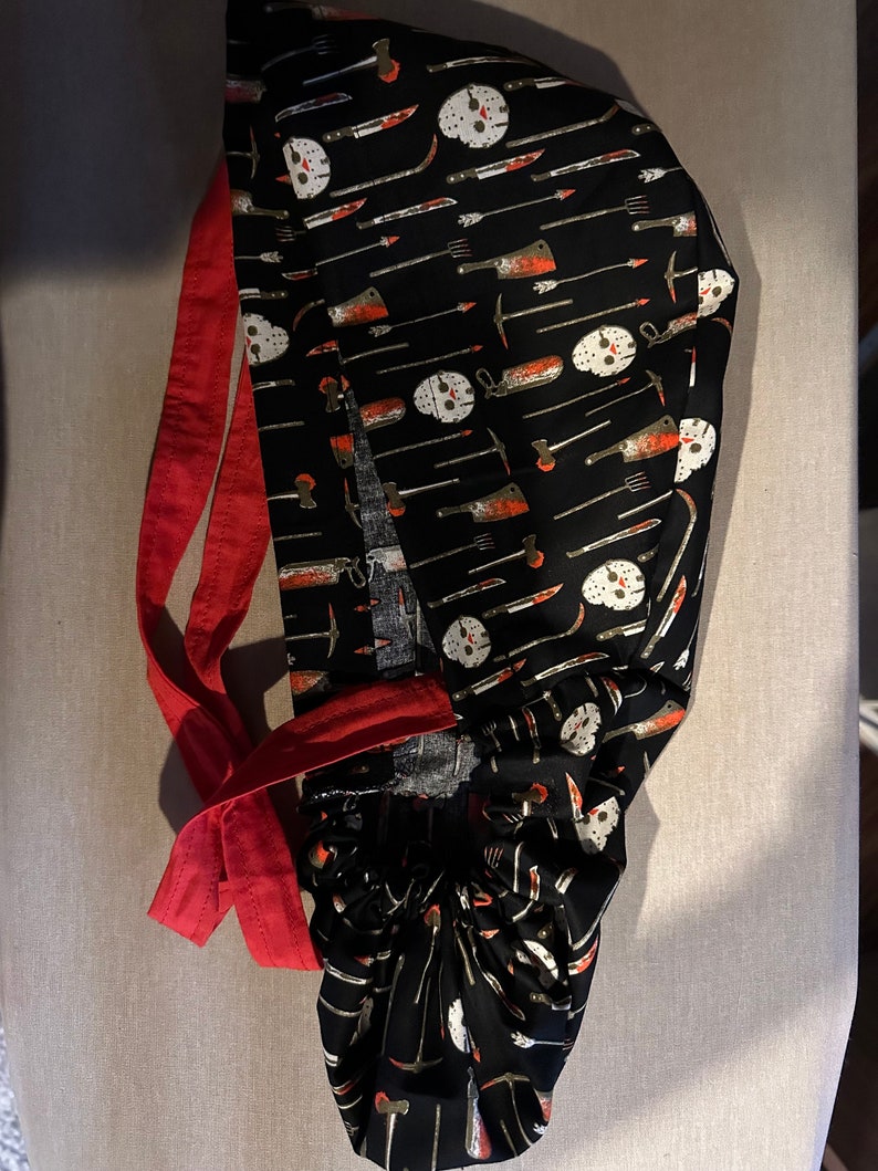 K&ouml;nnte beinhalten: Eine schwarze OP-M&uuml;tze mit roter Schleife. Die M&uuml;tze hat ein sich wiederholendes Muster aus wei&szlig;en Hockeymasken, Messern und anderen Waffen.