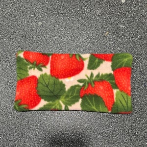 May include: A rectangular fleece item featuring a repeating pattern of bright red strawberries with green leaves and white flowers on a light pink background. The item is likely a small pillow or eye mask.