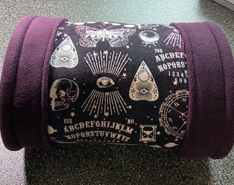 May include: A black and white patterned cylindrical pillow with a purple fleece exterior. The pattern features a Ouija board, a skull, an eye, a butterfly, and a crescent moon.