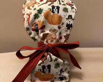 May include: A white fabric scrub hat with a pattern of dogs dressed in fall-themed costumes, including a pumpkin, a pilgrim hat, and a witch hat. The hat is tied with a burgundy ribbon.