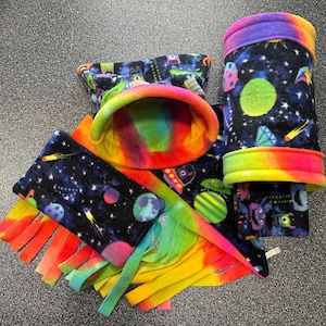 May include: A set of colorful fleece toys for small animals, featuring a rainbow tie-dye pattern, a space-themed print with planets, stars, and comets, and a monster print. The set includes a hideaway, a tunnel, and a play mat.