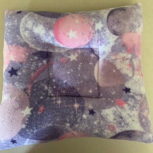 May include: A purple and pink pillow with a white, pink, and purple galaxy print. The pillow has a square opening in the center.
