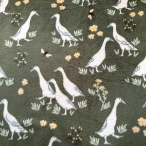 May include: A green fabric with a repeating pattern of white geese and yellow flowers. The geese are walking in a line and the flowers are scattered throughout the fabric.