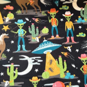 May include: A black fabric with a repeating pattern of green aliens riding horses, flying in spaceships, and wearing cowboy hats. The background features stars, planets, cacti, and skulls.