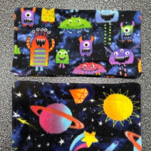 May include: Two rectangular fleece fabric squares. The top square has a black background with a colorful pattern of cartoon monsters. The bottom square has a black background with a colorful pattern of stars, planets, and a sun.