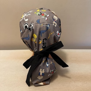 May include: Gray surgical scrub hat with a black tie. The hat features a cartoon print of characters from the Looney Tunes animated series.