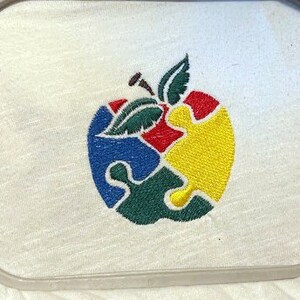 May include: An embroidered apple design with a green stem and leaves. The apple is made up of four puzzle pieces in red, blue, green, and yellow.