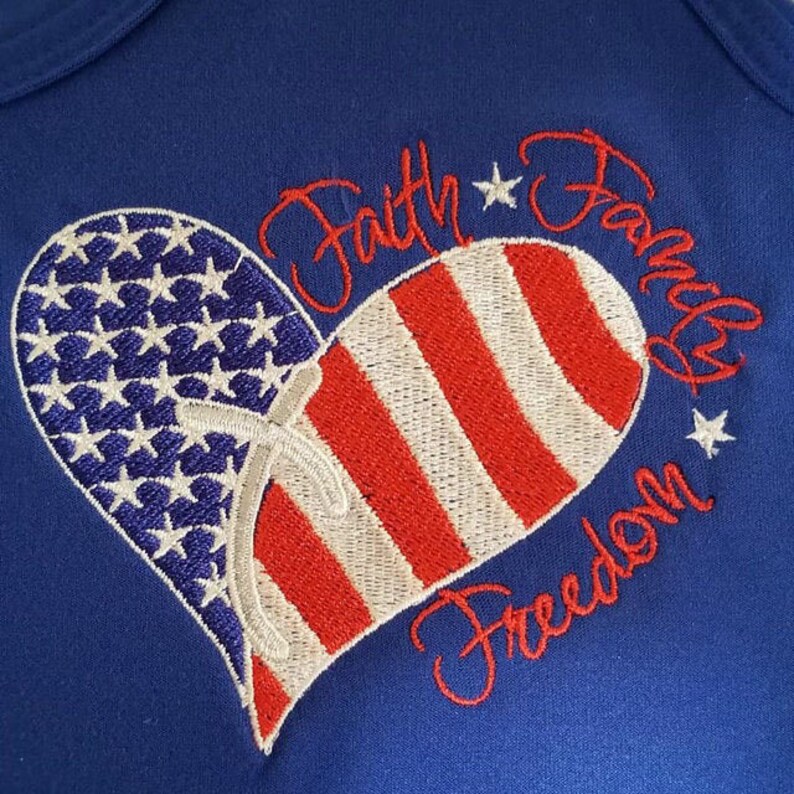 May include: Embroidered heart-shaped American flag design on a blue fabric background. The text "Faith Family Freedom" is stitched around the heart.