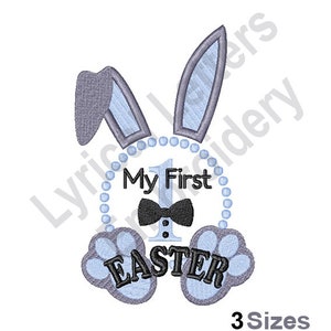 Könnte beinhalten: Stickerei eines blauen Hasen mit grauen Ohren und einer Fliege. Der Hase hält ein Schild mit der Aufschrift "Mein erstes Ostern" und der Zahl 1 in der Mitte.