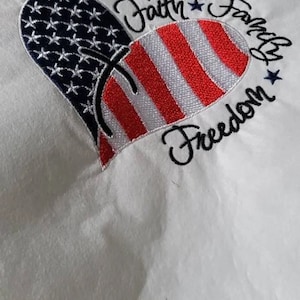May include: White fabric with an embroidered red, white, and blue heart. The heart is made up of an American flag design. The text "Faith Family Freedom" is embroidered around the heart.