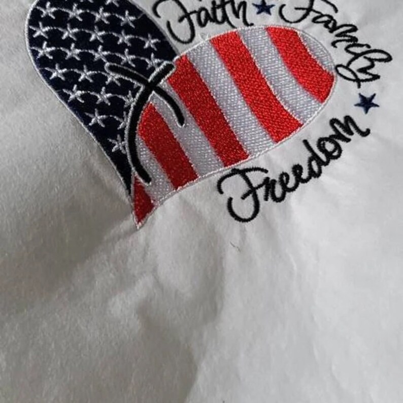 May include: White fabric with an embroidered red, white, and blue heart. The heart is made up of an American flag design. The text "Faith Family Freedom" is embroidered around the heart.