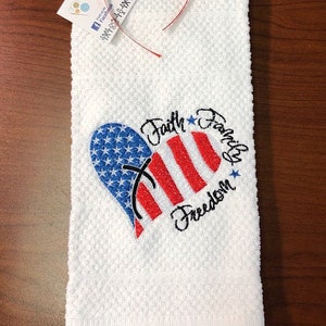 May include: White kitchen towel with an embroidered red, white, and blue heart with a cross in the center. The text "Faith Family Freedom" is embroidered around the heart.