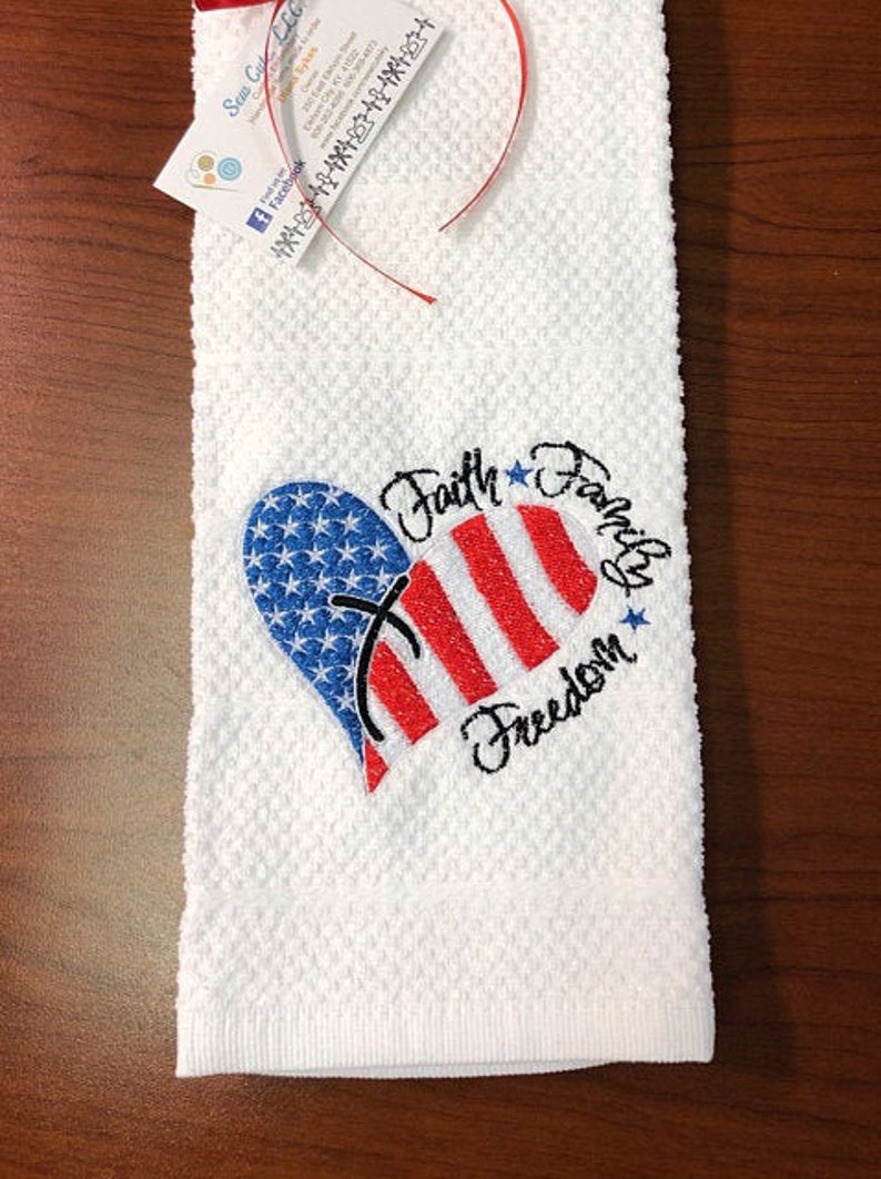 May include: White kitchen towel with an embroidered red, white, and blue heart with a cross in the center. The text "Faith Family Freedom" is embroidered around the heart.