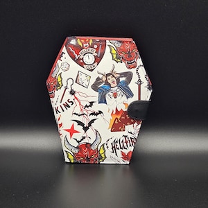 May include: A coffin-shaped wallet with a black snap closure. The white surface is covered in red and black illustrations, including a devil, bats, dice, and the words "Hellfire Club". A distressed image of a person is also featured.